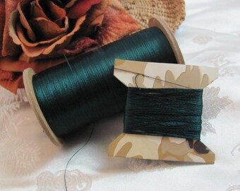May include: Two spools of dark green thread. One spool is on a wooden bobbin and the other is on a cardboard bobbin with a camouflage pattern.