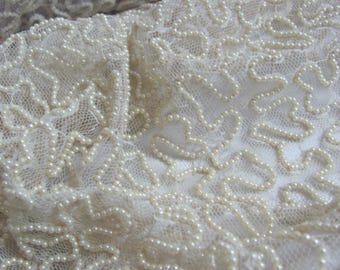 May include: Close-up of off-white lace fabric with a delicate pattern of small, creamy-colored pearls. The pearls are arranged in a flowing, organic design, creating a textured surface. The lace has a fine, open weave.