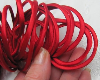 May include: Close-up of a bundle of bright red elastic cord, tightly coiled. The cord appears to be made of a smooth, satin-like material. The image shows a hand holding a portion of the cord, highlighting its texture and color.