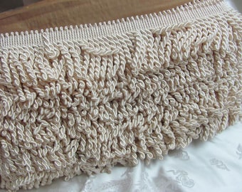 May include: Close-up of a cream-colored decorative fringe trim. The trim features a series of looped, twisted cords, creating a textured, voluminous effect. The top edge has a neat, corded border. This trim is suitable for various craft and home decor projects.