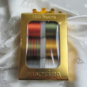 May include: A gold-colored box of Madeira thread spools. The box is labeled "100 Years" and features a clear window displaying various colored threads, including orange, green, and gold. The Madeira logo is visible on the box.