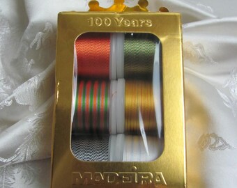 Puede incluir: Una caja dorada de carretes de hilo Madeira. La caja lleva la inscripción "100 Years" y presenta una ventana transparente que muestra hilos de varios colores, como naranja, verde y dorado. El logotipo de Madeira es visible en la caja.