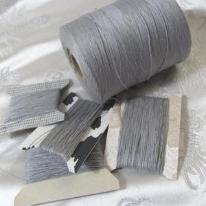 May include: A collection of gray thread spools and a large spool of thread. The spools vary in size and shape, some with decorative paper labels. The thread appears to be a light gray color, suitable for sewing or crafting projects.
