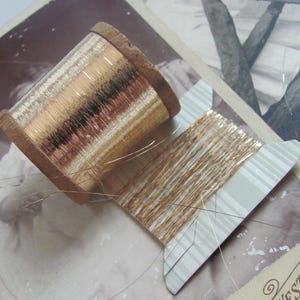May include: A spool of gold metallic thread wound around a wooden bobbin. The thread is wrapped around a cardboard spool with a white and grey striped pattern.