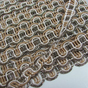 May include: A close-up of a decorative trim with a repeating pattern of interlocking loops. The trim is made of a white and gold fabric with a silver metallic thread woven throughout.