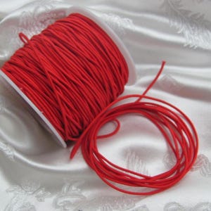 Könnte beinhalten: Eine Spule mit leuchtend rotem Gummiband liegt neben einer losen Spule aus demselben Material. Das Band ist eng auf eine weiße Kunststoffspule gewickelt. Das elastische Band hat eine leuchtend rote Farbe und eignet sich für verschiedene Bastel- und Nähprojekte.