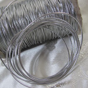 May include: A spool of silver metallic cord, with several strands unwound. The cord is tightly wound on a cardboard spool. The metallic cord is a bright silver color and appears to be flexible.
