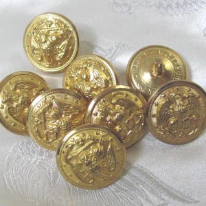 Puede incluir: Un conjunto de ocho botones redondos de metal de color dorado. Cada botón presenta un diseño en relieve de un águila con las alas extendidas, rodeado por un borde de pequeñas estrellas. La superficie de los botones tiene un acabado pulido.