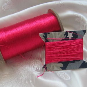 May include: Two spools of bright pink thread. One spool is large and cylindrical, while the other is smaller and has a camouflage pattern on the sides. The thread is tightly wound on the spools, ready for use in sewing or crafting projects.