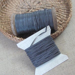May include: A dark grey metallic cord wrapped around a cylindrical wooden spool and a flat white cardboard bobbin. The cord features a fine, shimmering texture and is wound tightly in multiple layers across both the spool and the bobbin.