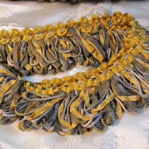 May include: Close-up of decorative trim featuring a combination of gold and gray colors. The trim has a looped fringe design with a woven texture. The trim is likely used for embellishing clothing or home decor items.