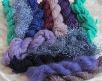 May include: A collection of yarn in various shades of blue, purple, and teal. The yarn is arranged in a pile on a white surface.