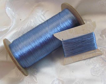 Könnte beinhalten: Eine Spule mit hellblauem Garn, das um eine Holzspule gewickelt ist. Das Garn ist glänzend und hat einen dezenten Schimmer. Die Spule liegt auf einer weißen Stoffoberfläche.