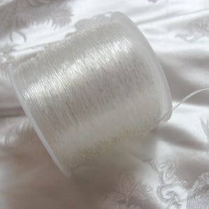 May include: A spool of clear, elastic beading thread. The thread is tightly wound inside a transparent plastic container. The thread is a light, translucent color, and a single strand extends from the spool.
