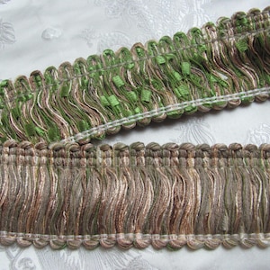 May include: Close-up of decorative fringe trim in shades of green, brown, and beige. The trim features a textured design with a scalloped edge and a woven top. Ideal for home decor and craft projects.