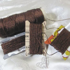 May include: Various spools of brown metallic thread are arranged on a white, textured surface. The thread is wound on cardboard bobbins and a large white spool. The thread has a shiny, reflective quality, suggesting it is made of a metallic material.