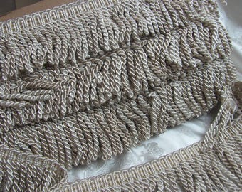 May include: Close-up of beige rope fringe trim. The trim features a twisted rope design with a lighter beige border. The fringe is densely packed, creating a textured and decorative element. This trim is suitable for various craft and design projects.