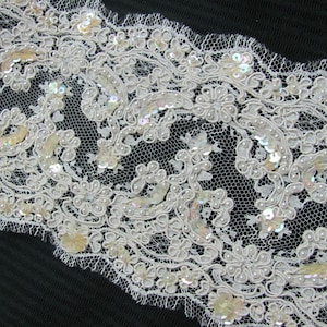 May include: Ivory lace trim with floral and scrollwork patterns. The lace is embellished with small, iridescent sequins and tiny, pearl-like beads. The trim has a scalloped edge and is suitable for various craft projects.