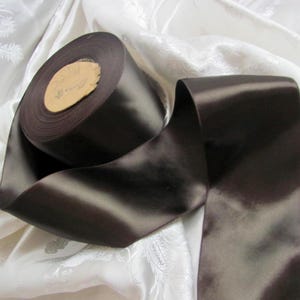 Könnte beinhalten: Eine Rolle dunkelbraunes Satinband mit glänzendem Glanz. Das Band ist abgerollt und zeigt eine Länge des gleichen Materials. Der Hintergrund ist ein weißer Stoff mit einem dezenten Muster, das einen Kontrast zum dunklen Band bildet.