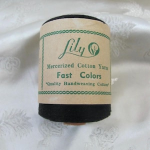 May include: A spool of black mercerised cotton yarn wrapped in a tan paper label. The label features green text that reads Lily, Mercerised Cotton Yarns, Fast Colours, and Quality Handweaving Cottons, framed by a decorative green rope-style border.
