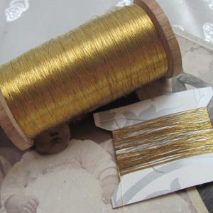 Könnte beinhalten: Eine Holzspule mit goldfarbenem Metallic-Garn und eine Karte mit demselben Garn werden gezeigt. Der Faden ist um eine Holzspule gewickelt. Die Karte ist weiß mit einem grau-weißen Muster. Der Hintergrund enthält ein Vintage-Foto.