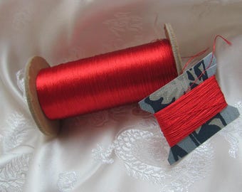 May include: Two spools of red thread. One is a large spool with a cardboard core, and the other is a smaller spool with a patterned cardboard core. The thread has a shiny, silky appearance. The spools are on a white, patterned fabric.