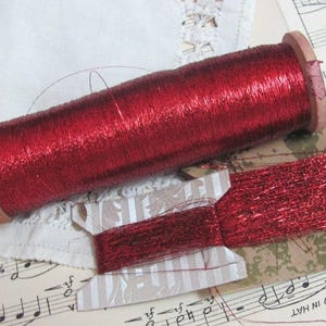 May include: A cylindrical wooden spool wrapped with vibrant metallic red thread, accompanied by a flat cardboard bobbin also wound with the same shiny red thread. The thread has a distinct reflective quality and a smooth, thin texture.
