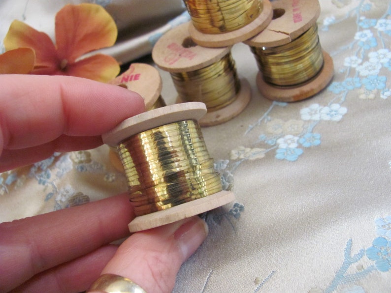 May include: Close-up of a spool of gold metallic thread wound around a wooden bobbin. The spool is being held in a hand.