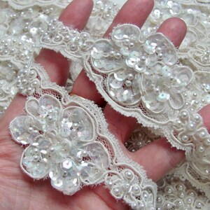 May include: Ivory lace trim with floral embellishments. The trim features delicate, embroidered flowers adorned with sequins and small, round pearls. The lace has a scalloped edge and is ideal for bridal or craft projects.