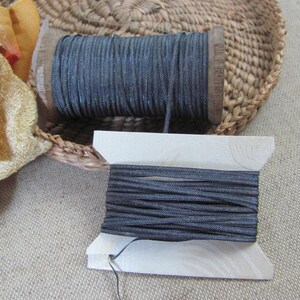 May include: A dark grey metallic cord wrapped around a wooden spool and a flat white cardboard bobbin. The cord has a subtle shimmering texture and is wound tightly in multiple horizontal layers across both the spool and the flat bobbin.