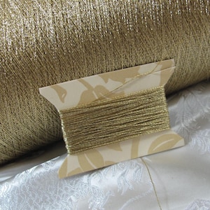 May include: A large spool of metallic gold thread positioned behind a smaller, flat cardboard bobbin wrapped with the same shimmering gold thread. The bobbin features a light cream-colored surface with a subtle, repeating leaf pattern printed on it.