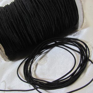 May include: A spool of black elastic cord sits next to a loose coil of the same material. The cord is tightly wound on a cardboard spool. The background is a white, textured fabric. The cord appears to be approximately 1/8 inch in diameter.