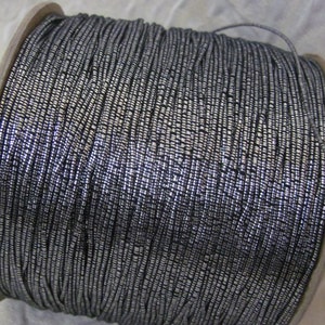 May include: A spool of silver metallic cord. The cord is wound tightly around the spool, creating a dense, textured surface.