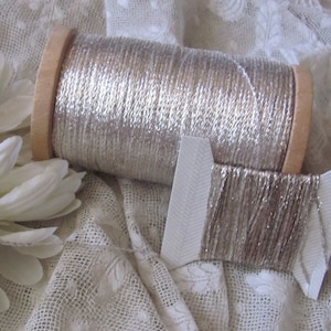 May include: A spool of silver metallic thread wound on a wooden bobbin. The thread is displayed on a white lace fabric background.