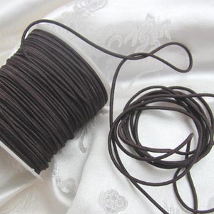 Puede incluir: Un carrete de cordón elástico marrón oscuro. El cordón está bien enrollado en un carrete de plástico blanco. Una longitud del cordón está desenrollada y enrollada sobre una superficie de tela blanca. El cordón parece liso y flexible, adecuado para varios proyectos de manualidades.