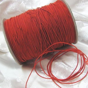 May include: A spool of bright red elastic cord. The cord is tightly wound around a cardboard core. A length of the same red cord is unwound and lies in a loose coil in front of the spool. The background is a white, textured fabric.