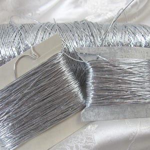 May include: A large black spool of metallic silver thread positioned behind two smaller cardboard bobbins, each wrapped with the same shiny silver thread. The thread has a reflective, smooth texture and is wound tightly around the cylindrical and flat supports.
