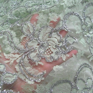 May include: Close-up of a delicate lace fabric in a soft green hue, adorned with intricate floral patterns and shimmering silver accents. The lace is held up, revealing its fine texture and design, perfect for apparel or crafts.