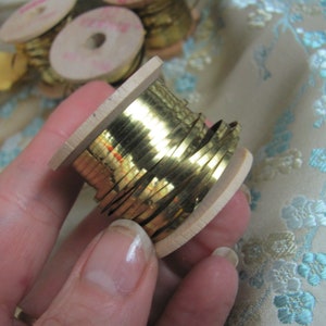 May include: A spool of gold metallic trim wound around a wooden bobbin. The trim is shiny and has a slightly textured surface.