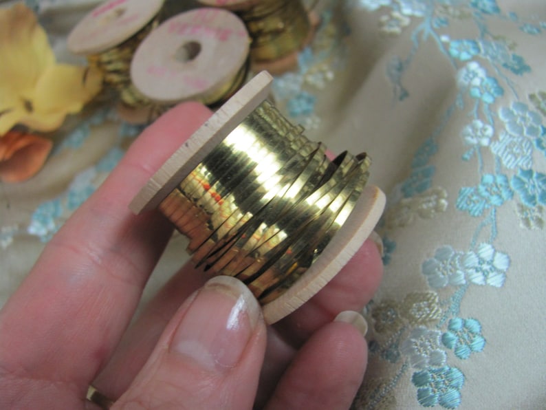 May include: A spool of gold metallic trim wound around a wooden bobbin. The trim is shiny and has a slightly textured surface.