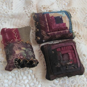 May include: Three handmade fabric pincushions in shades of brown, burgundy, and cream. The pincushions are made from patchwork fabric and have a quilted design.