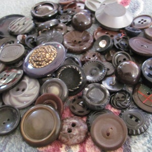 May include: A collection of vintage buttons in various shades of brown, black, and grey. The buttons are made of different materials, including plastic, metal, and wood. Some buttons have intricate designs, while others are plain.