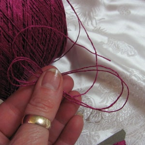 May include: A close-up of a spool of deep pink yarn with a strand of yarn being held by a finger. The yarn is smooth and has a slight sheen.