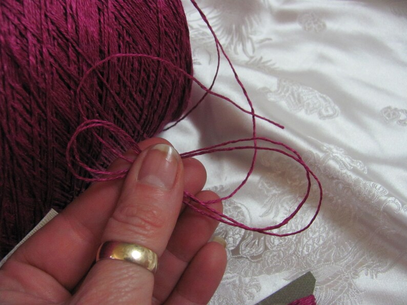 May include: A close-up of a spool of deep pink yarn with a strand of yarn being held by a finger. The yarn is smooth and has a slight sheen.