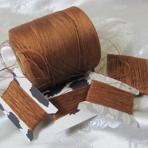May include: A large spool of brown thread sits next to several smaller spools. The thread is a rich, warm brown color. The smaller spools have decorative paper labels with black and white cow print and floral patterns.