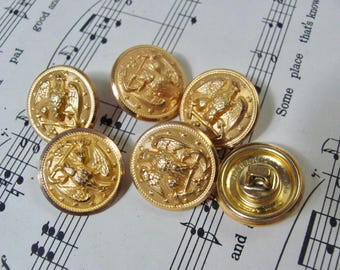 May include: A collection of six round gold-toned metal buttons, each featuring a raised eagle emblem surrounded by small stars and a textured border. One button is flipped over to reveal a metal shank attachment on the back surface.