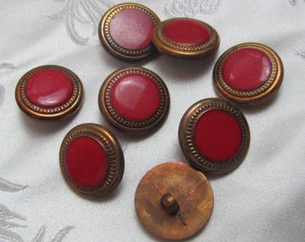 May include: Seven red and gold buttons with a raised rim and a smooth, shiny surface. The buttons are arranged in a semi-circle on a white fabric background.