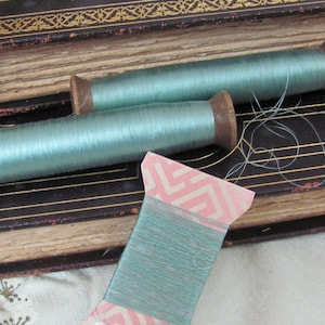 May include: Three spools of teal blue thread, two on wooden spools and one on a pink and white spool. The spools are sitting on top of an old, leather-bound book.