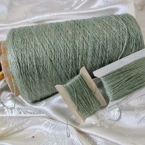 May include: A large sage green yarn cone accompanied by two smaller wooden spools wrapped in the same sage green yarn. The yarn has a textured, multi-ply appearance, and the spools are positioned in front of the main cone to display the thread.