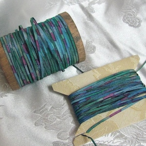 May include: Two spools of thin, flat ribbon featuring a repeating pattern of teal, dark blue, and magenta vertical segments. One spool is made of light-colored wood, while the other is a flat, rectangular piece of cream-colored cardstock.